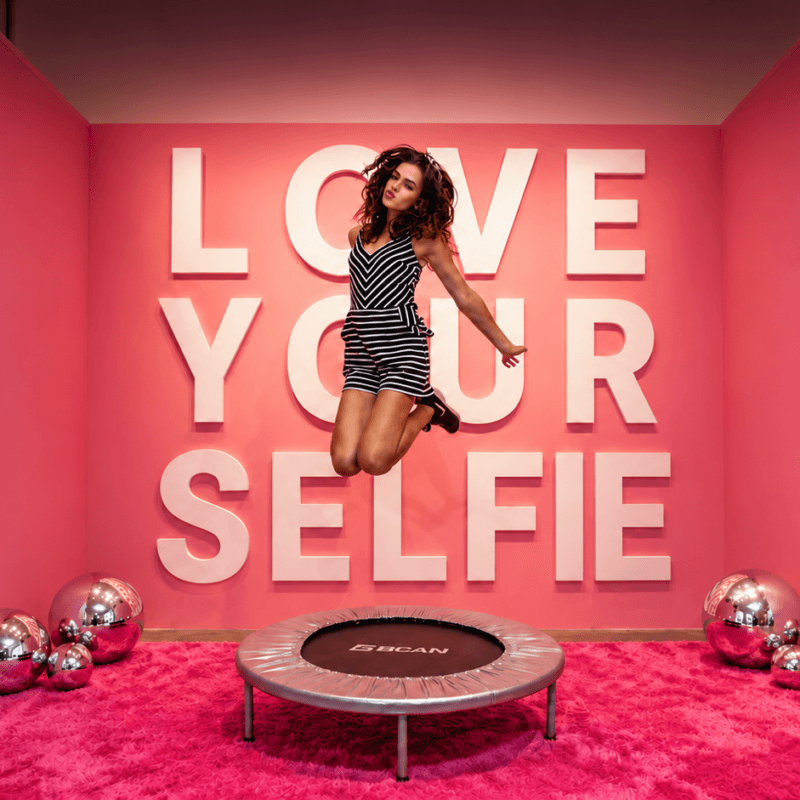 Woman jumping in front of a pink LOVE YOURSELFIE photo backdrop