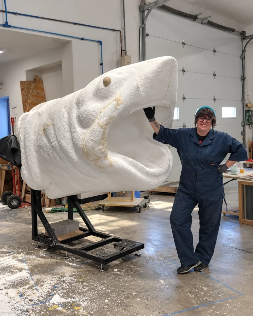 Person standing beside an oversized white foam shark head sculpture in a workshop, showing its scale